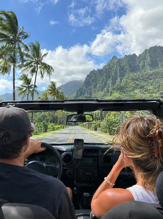 View from the back seat of an open-top car on a palm-lined road toward green mountains