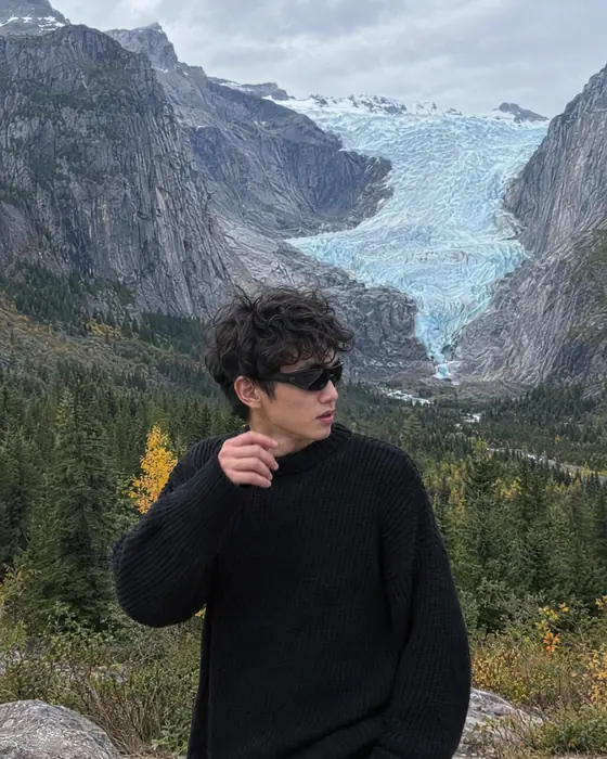 A young man in a black sweater and sunglasses standing above a valley, a glacier behind him