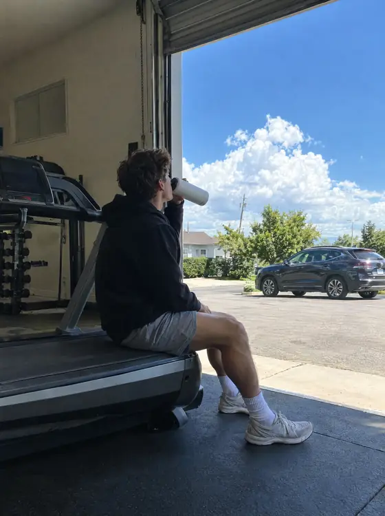 A man drinking from a water bottle, sitting on a treadmill by an open garage door on a sunny morning